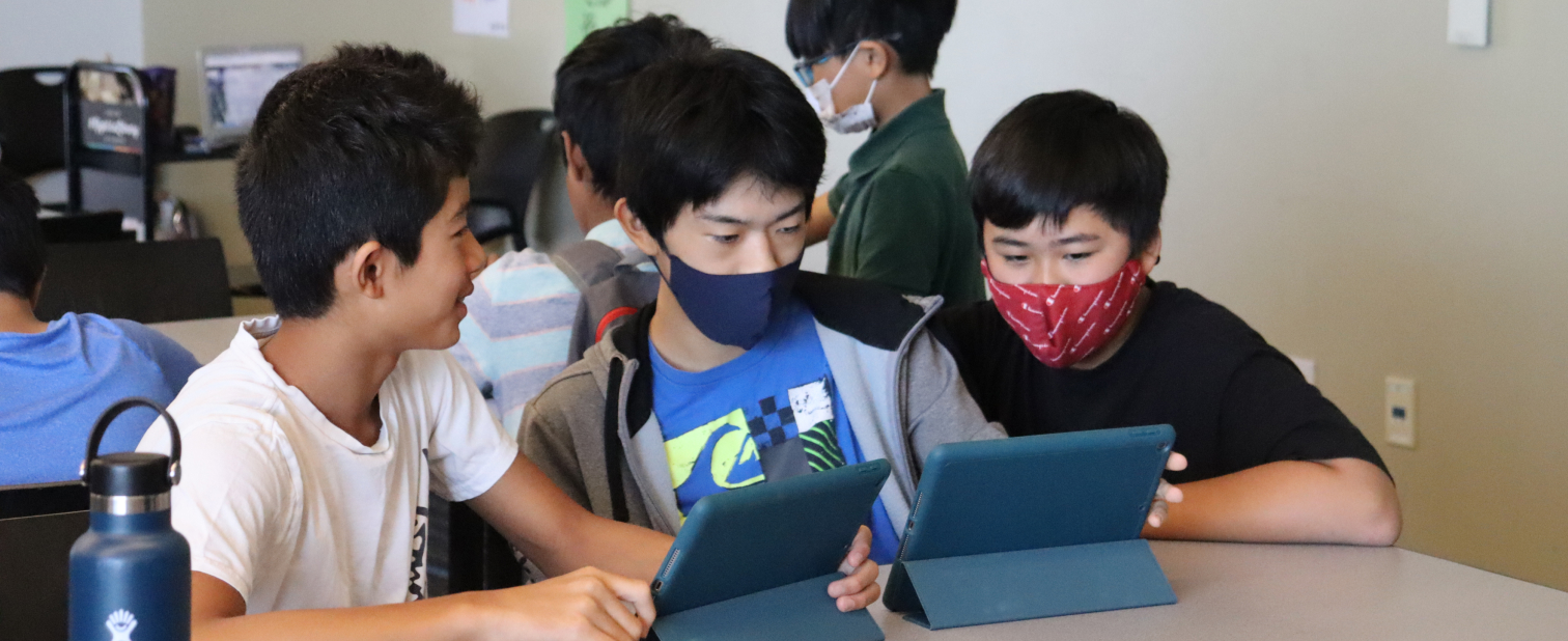 Three boys use iPads. One is smiling, the other two wear face masks.