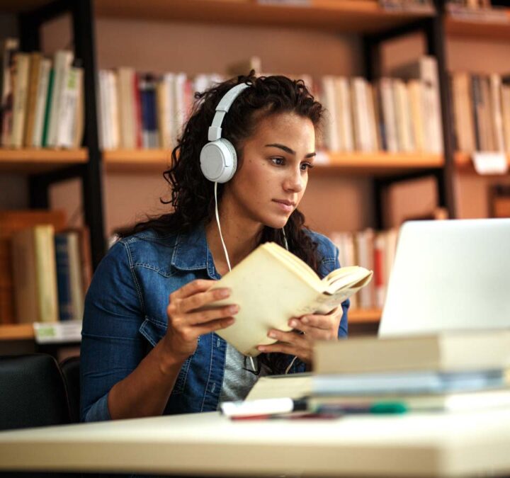 A woman with headphones looking at the computer while holding a book.