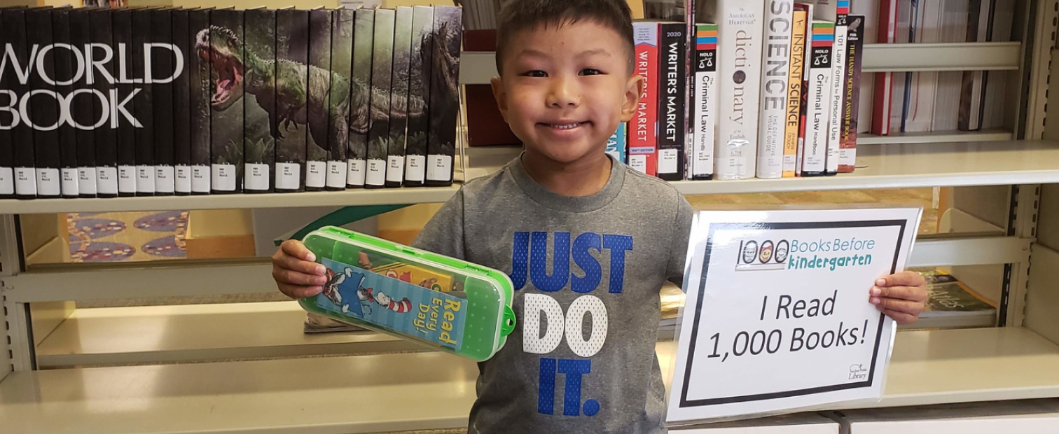 A young boy smiles while holding a pencil case and a sign reading 'I Read 1,000 Books!'