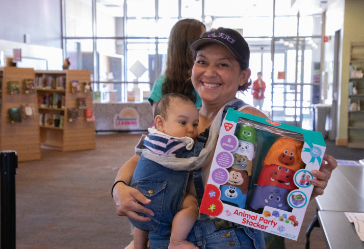 A woman smiles at the camera while holding a baby and a new toy.