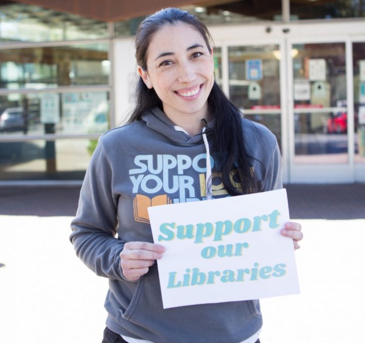 A young woman smiles at the camera while holding a sign that reads, 'Support our Libraries'.