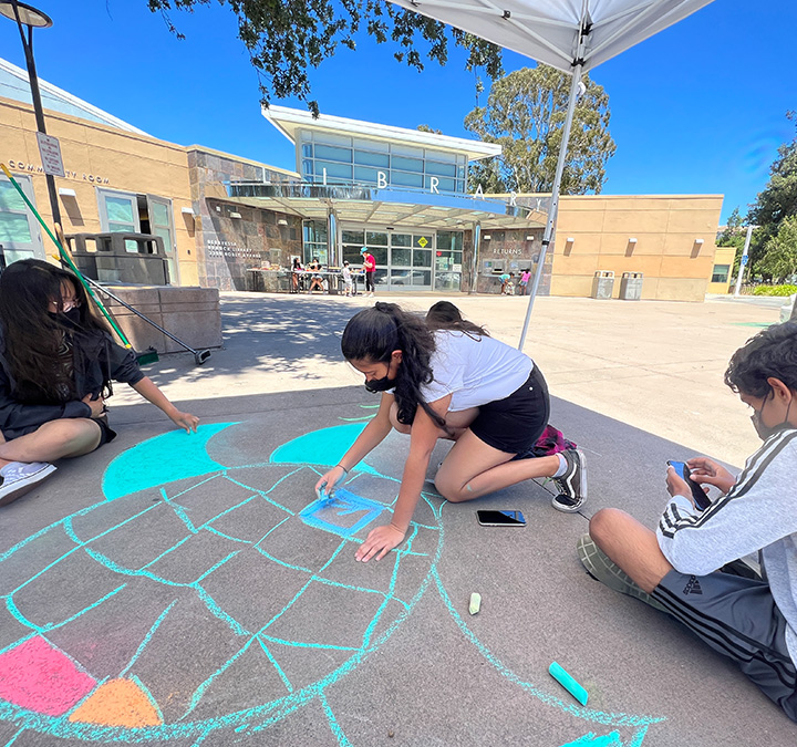 Young people coloring with chalk on the sidewalks outside a library branch.